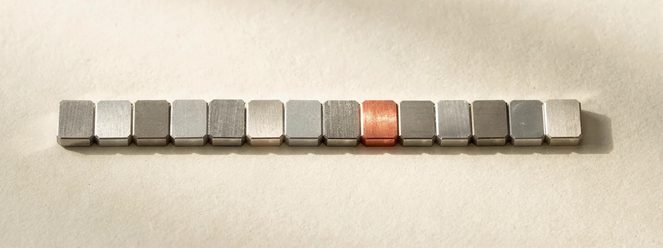 A row of machined steel blanks of the same shape, one in copper, on paper.