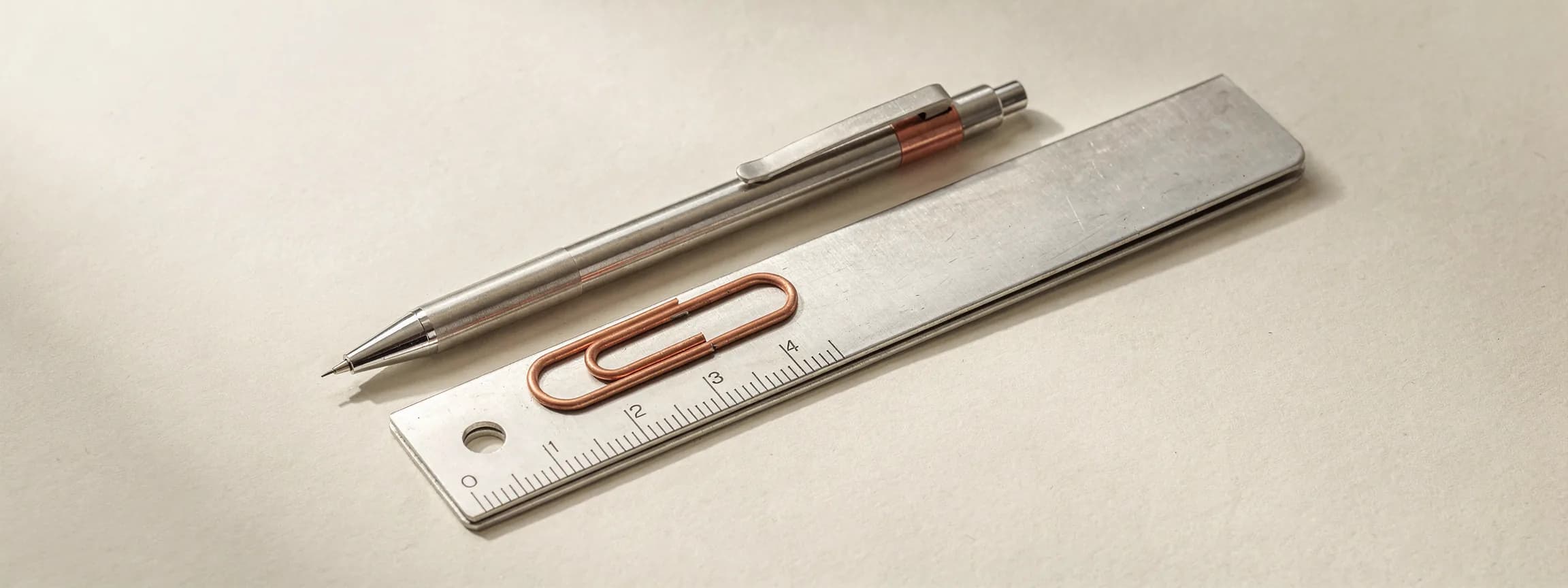 A stainless mechanical pencil and a copper paper clip resting on a folded steel rule, on paper.