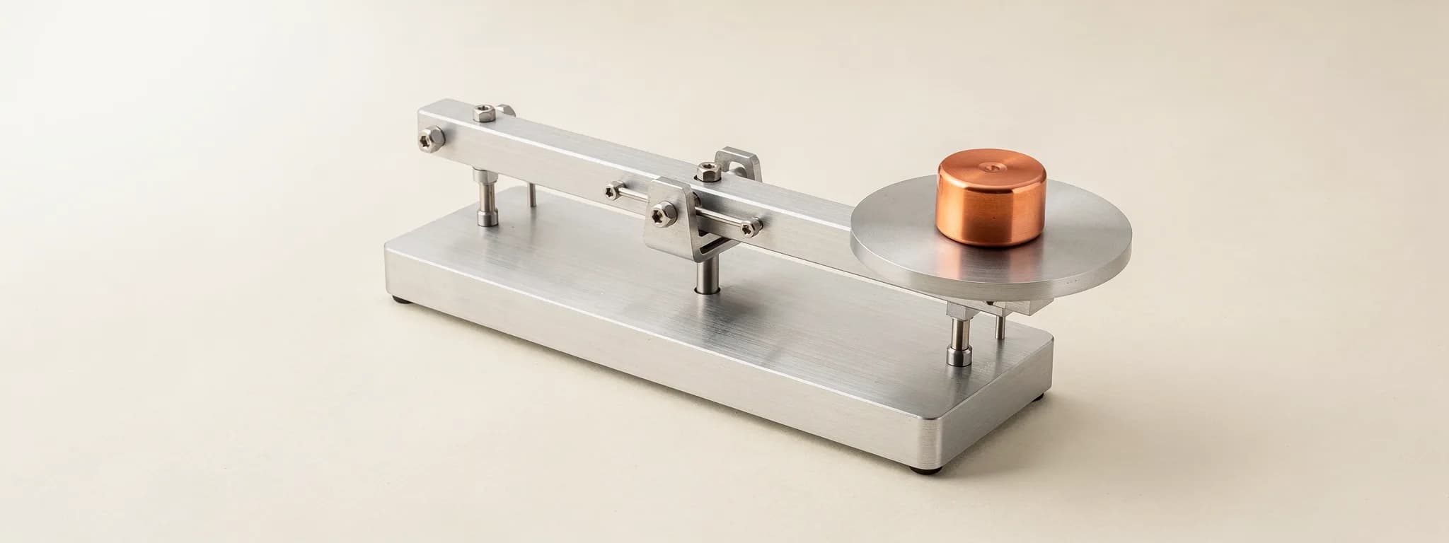 A brushed aluminium balance on paper, tipped by a single copper weight on its pan.